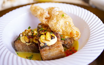 A delicately plated dish shows meat topped with corn. 