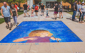 A crowd walks around a sidewalk chalk mural depciting a young, blonde-haired girl.